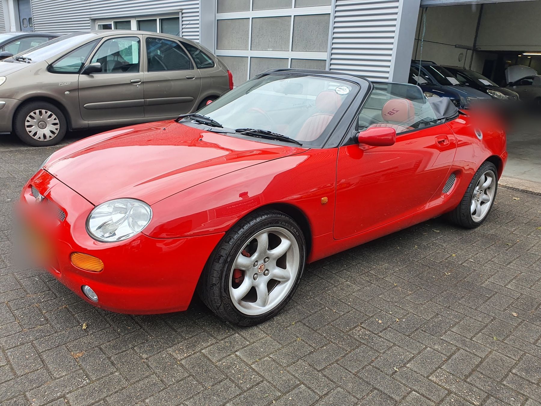 Rode MG F cabrio op het terrein, met auto's in de werkplaats erachter
